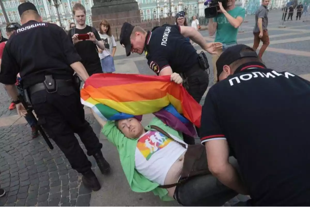 Un grupo de policías detiene a una persona que sostiene una bandera arcoíris mientras varios transeúntes observan y algunos toman fotos.