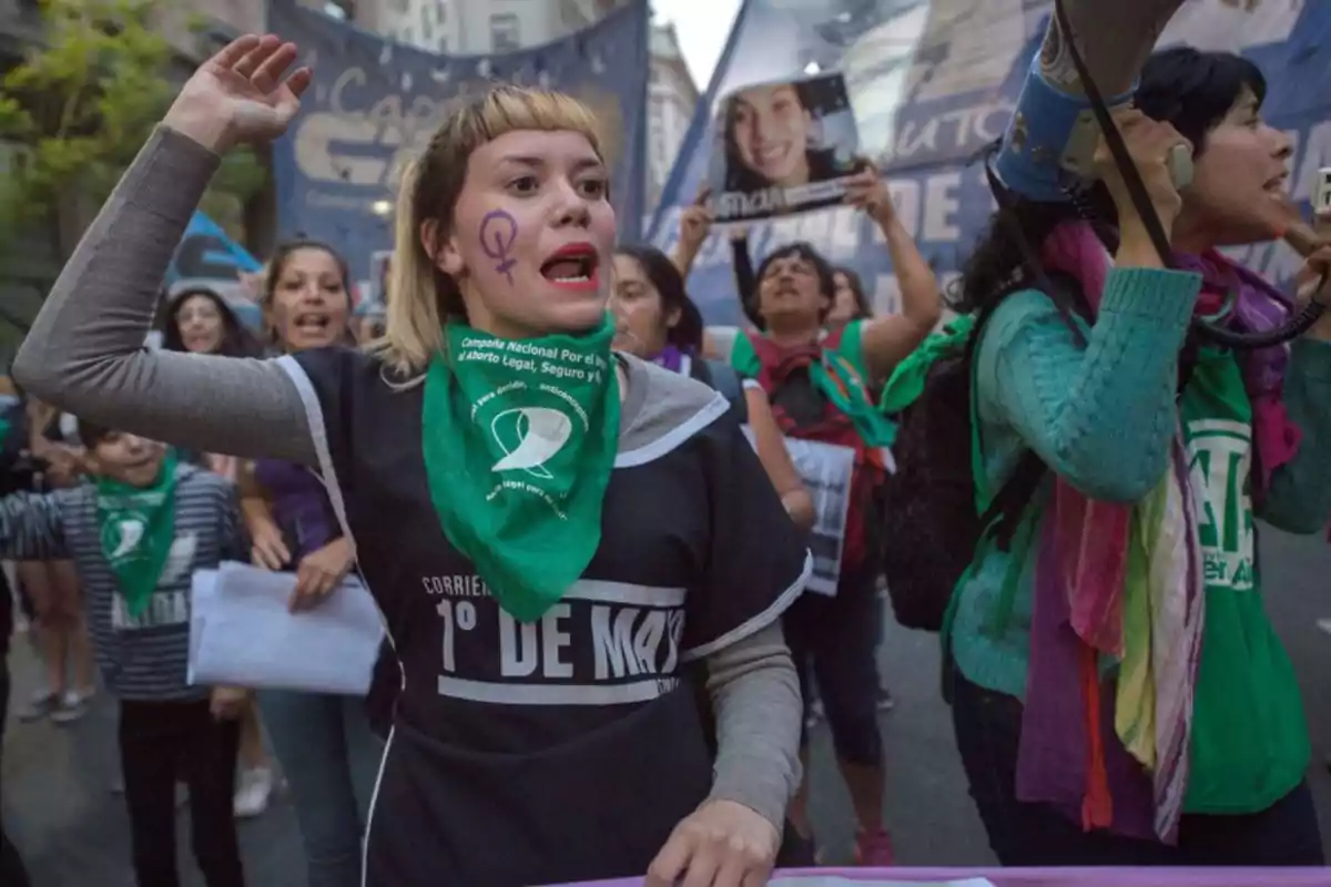 Un grupo de personas participa en una manifestación, algunas llevan pañuelos verdes y una mujer tiene un símbolo feminista pintado en la cara.