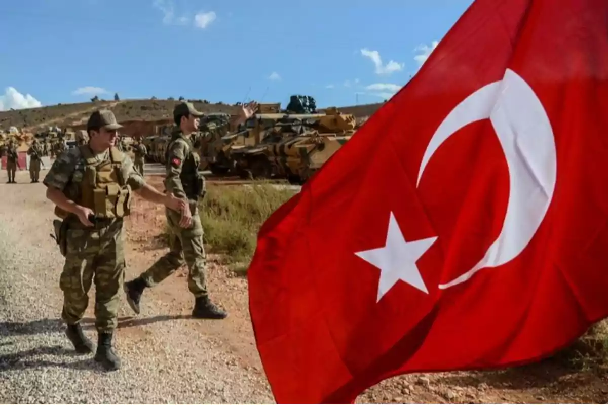 Soldados en uniforme camuflado caminan junto a una bandera de Turquía ondeando al viento con vehículos militares al fondo en un paisaje árido.