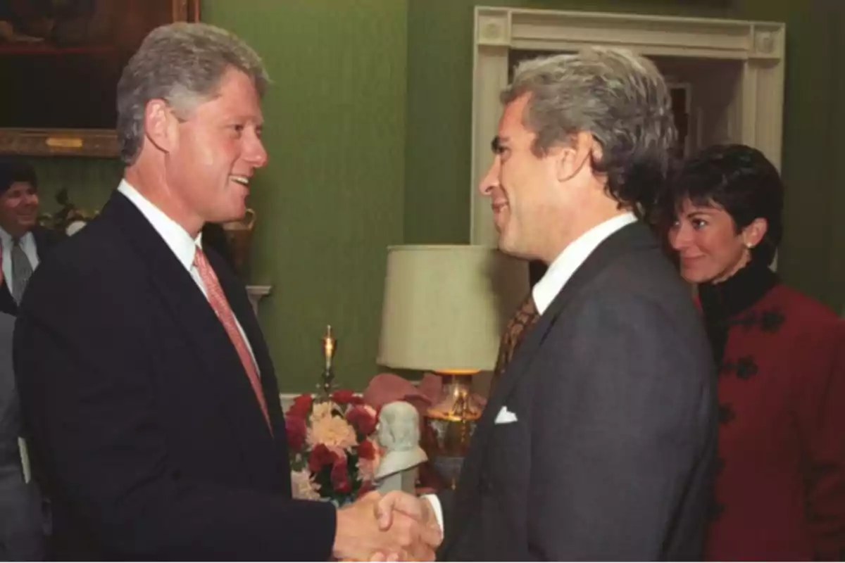 Two men in suits shake hands in a room decorated with flowers and a lamp, while a woman watches in the background.