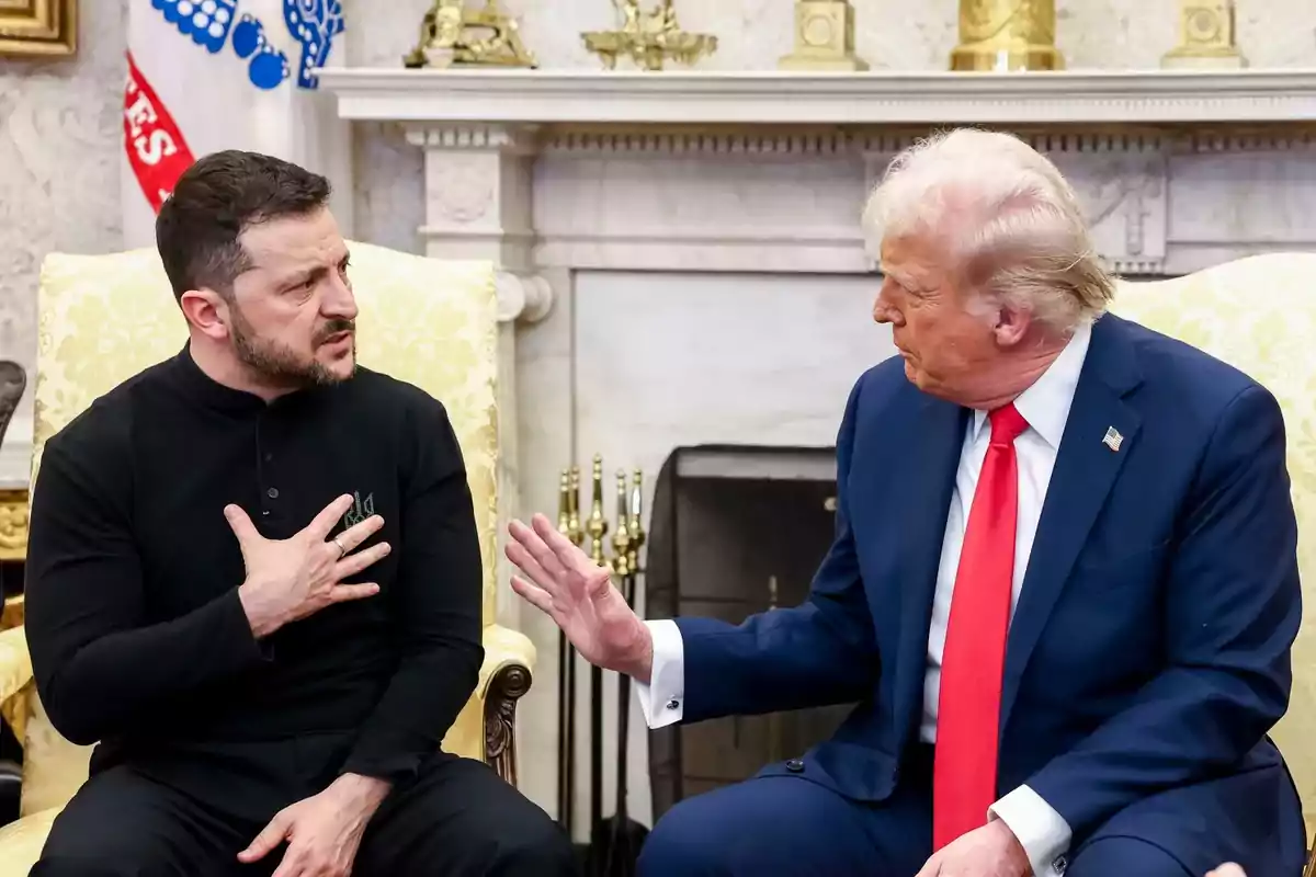 Two men sitting in armchairs, one in a blue suit and red tie, the other in a black jersey, talking in a room with a fireplace and golden decorations.