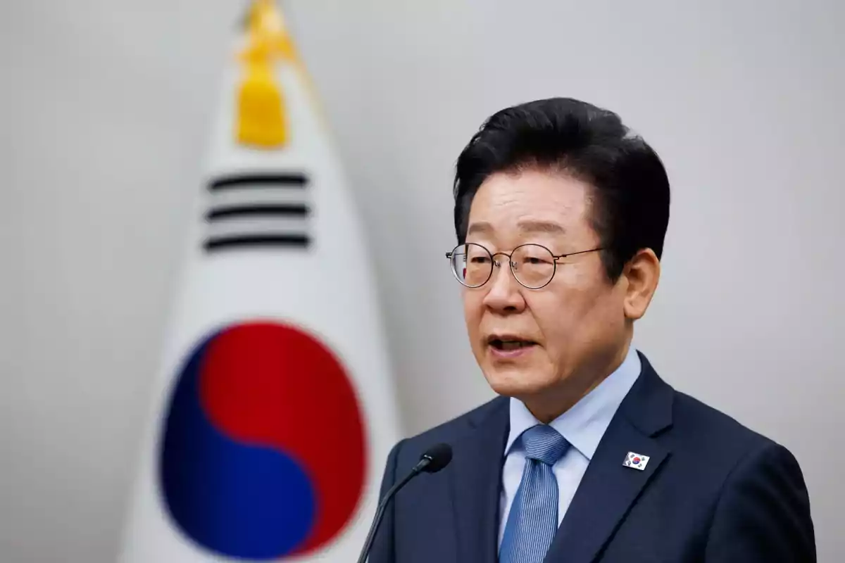 A man in glasses and a blue suit speaks in front of a microphone with the South Korean flag in the background. A man in glasses and a blue suit speaks in front of a microphone with the South Korean flag in the background.