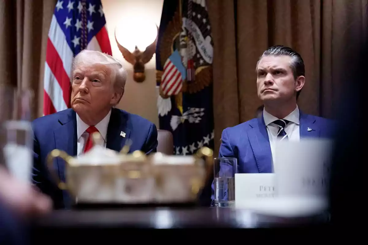 Dos hombres en traje sentados en una mesa con banderas estadounidenses y un emblema presidencial de fondo. Trump y Hegseth