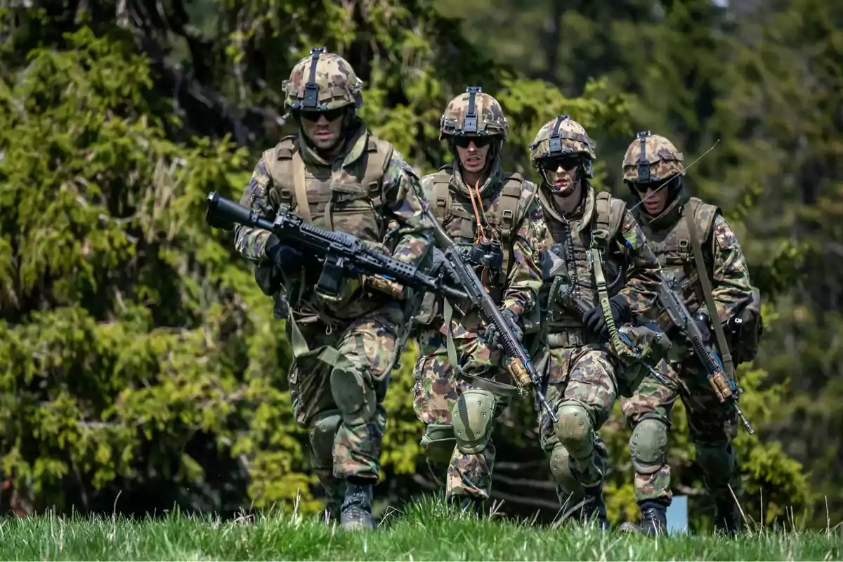 Four soldiers in camouflage uniforms and military gear move together across a green field surrounded by trees.
