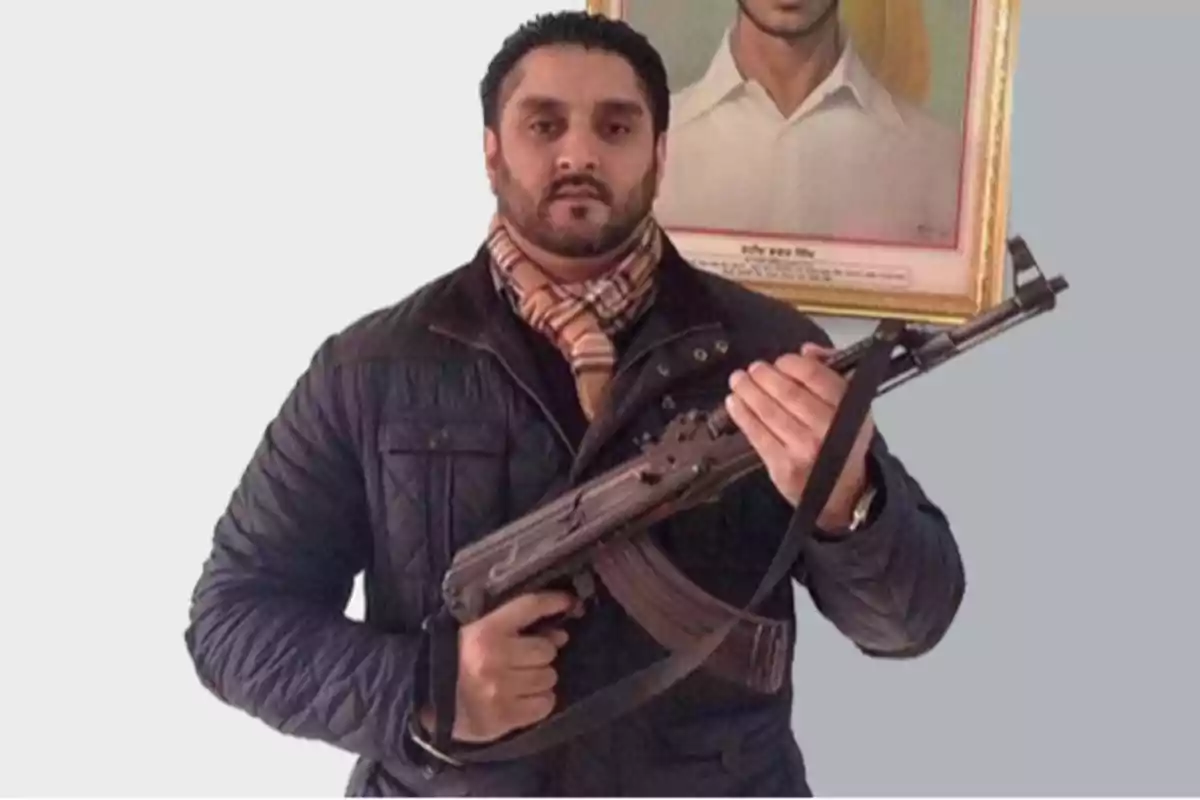 A man in a dark jacket and scarf holds a rifle in front of a framed portrait.