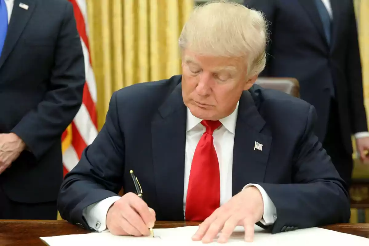 A man in a dark suit and red tie is signing a document at a desk.