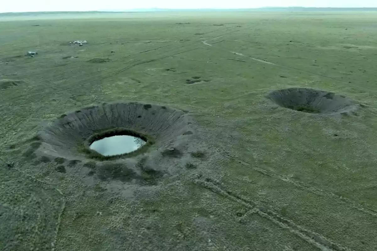 Vista aérea de un paisaje con dos grandes cráteres en el suelo, uno de ellos lleno de agua, rodeados de vegetación y caminos de tierra.