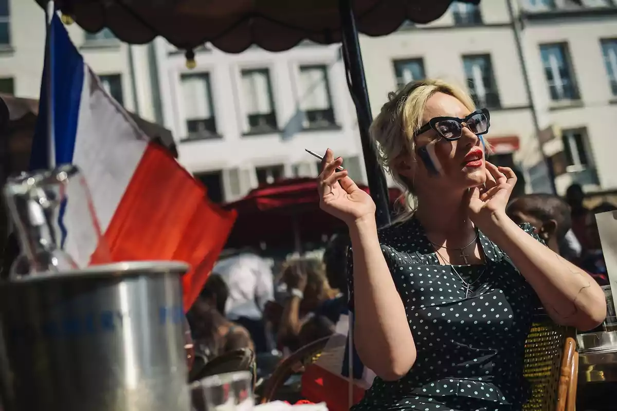 Mujer con gafas de sol y vestido de lunares sentada en una terraza, con la cara pintada con los colores de la bandera francesa y una bandera de Francia al fondo Mujer con gafas de sol y vestido de lunares sentada en una terraza, con la cara pintada con los colores de la bandera francesa y una bandera de Francia al fondo