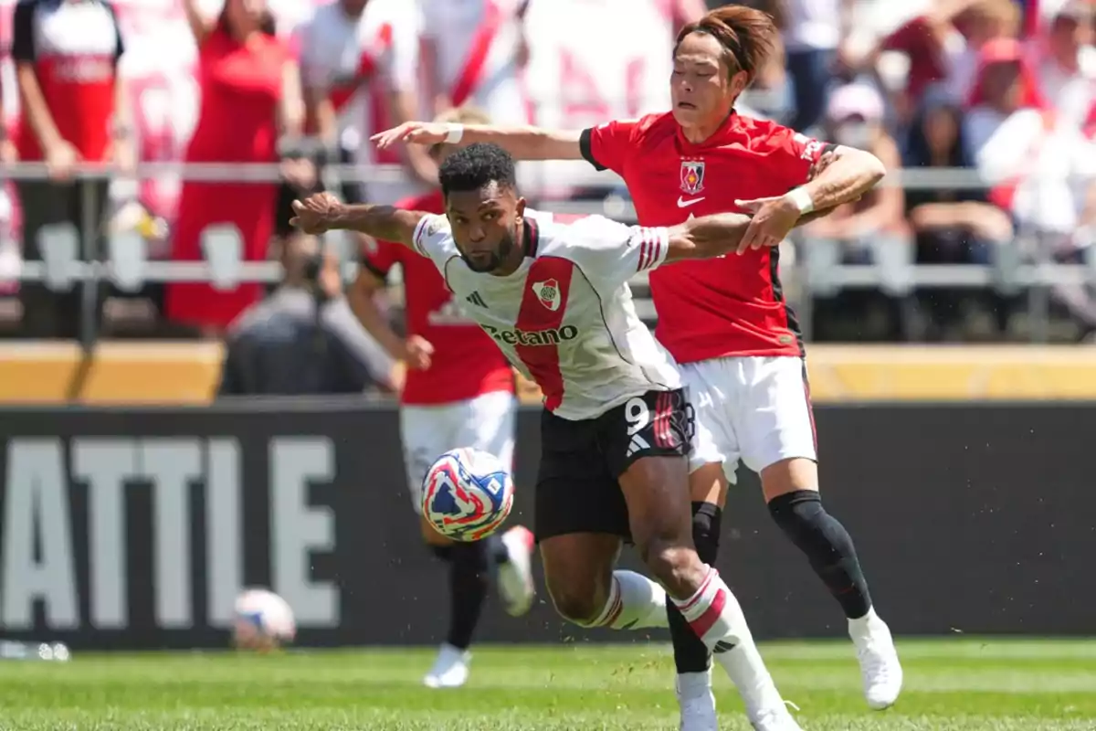 Two players compete for the ball during a match, one wearing a white uniform and the other wearing a red uniform, while the crowd watches from the stands. Two players compete for the ball during a match, one wearing a white uniform and the other wearing a red uniform, while the crowd watches from the stands.