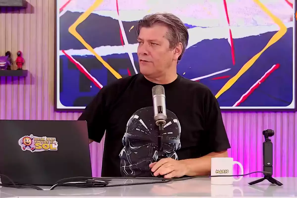 Gray-haired man sitting in front of a microphone with a laptop and a white mug on the table, with an abstract painting and a light-colored paneled wall in the background. Gray-haired man sitting in front of a microphone with a laptop and a white mug on the table, with an abstract painting and a light-colored paneled wall in the background.