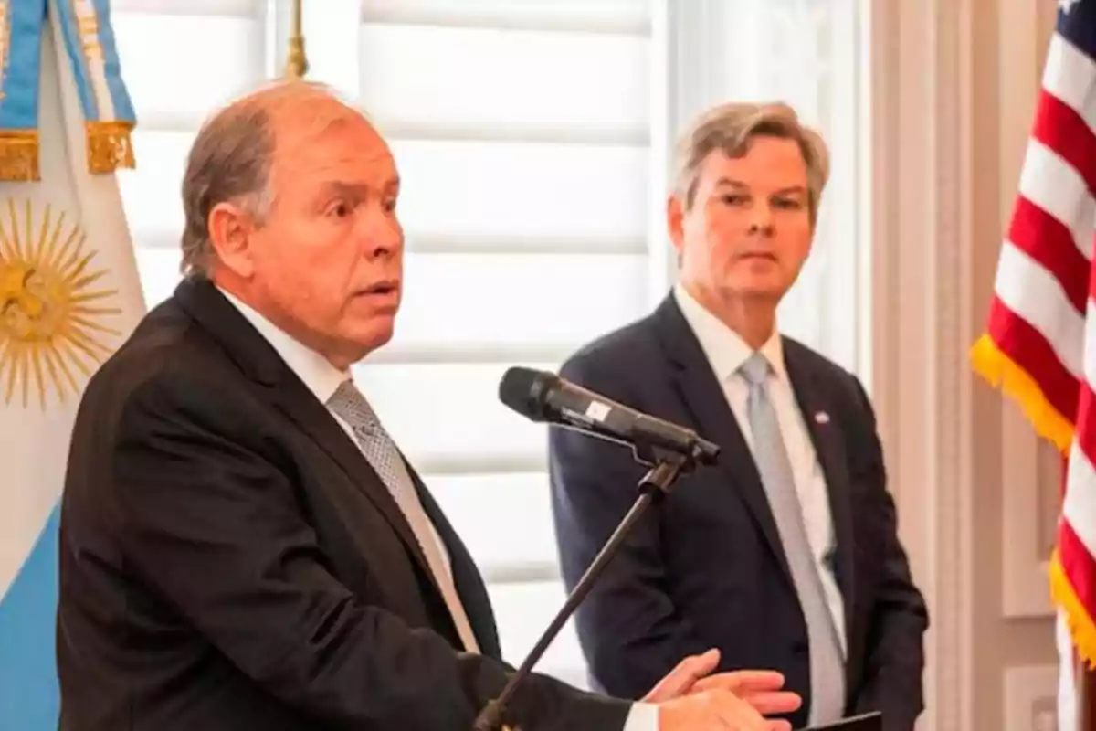 Dos hombres en trajes formales en un evento con banderas de Argentina y Estados Unidos.