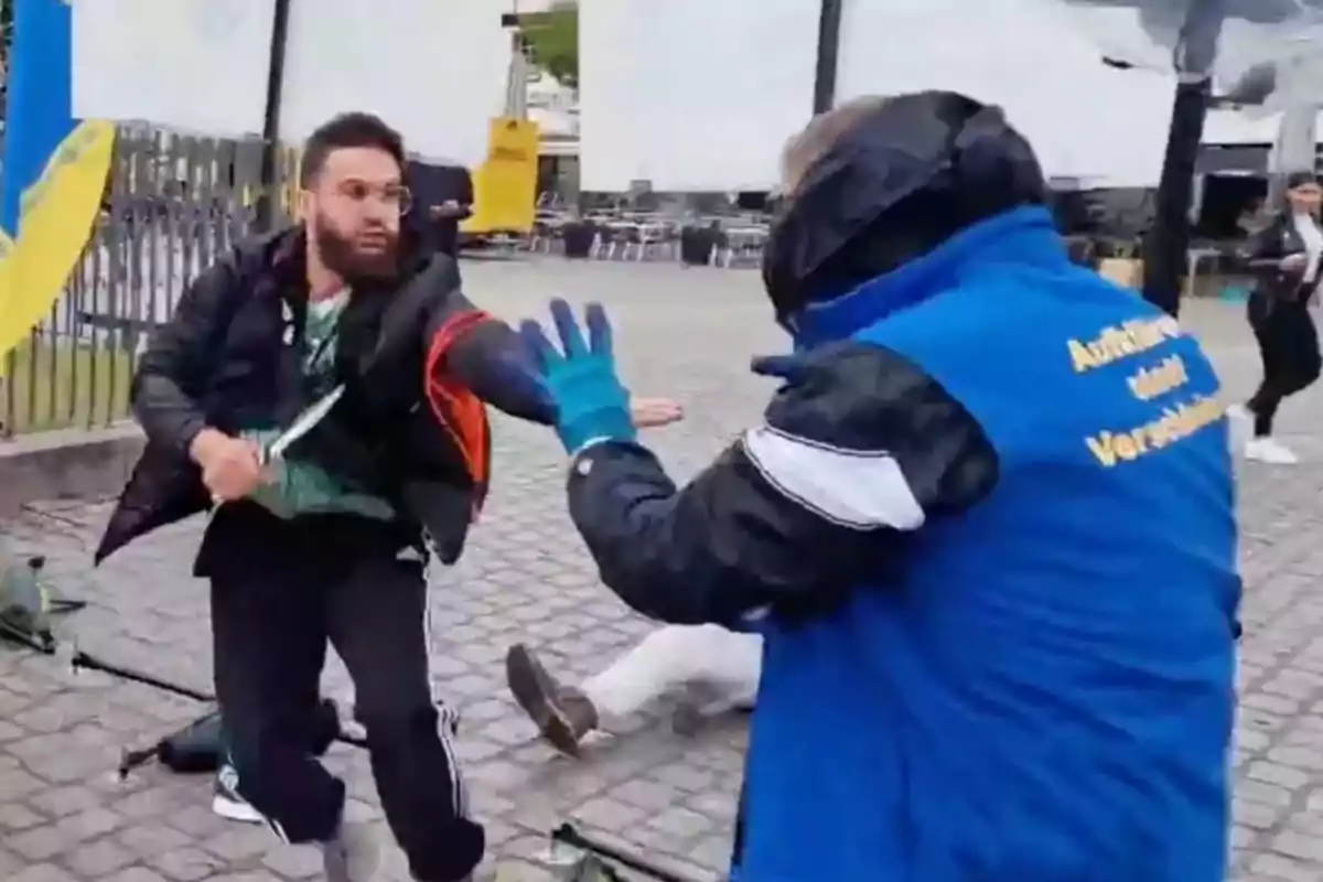 A bearded man holds a knife while another man in a blue jacket tries to stop him on a cobblestone street. A bearded man holds a knife while another man in a blue jacket tries to stop him on a cobblestone street.