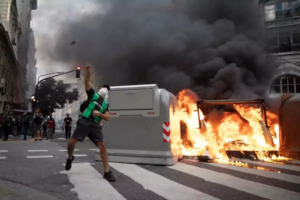 Un manifestante enmascarado lanza un objeto mientras un contenedor arde en llamas en una calle llena de humo.