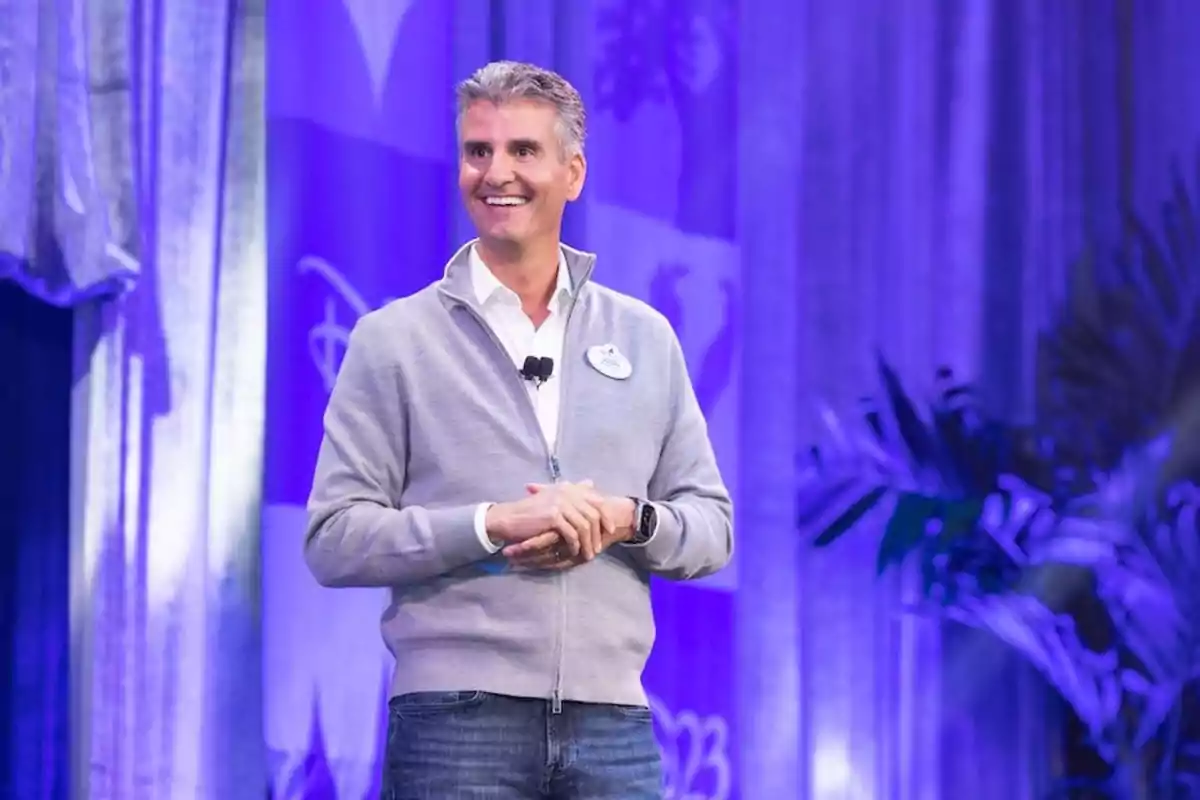 Un hombre sonriente con una chaqueta gris y jeans está de pie en un escenario con un fondo azul y plantas. Un hombre sonriente con una chaqueta gris y jeans está de pie en un escenario con un fondo azul y plantas.