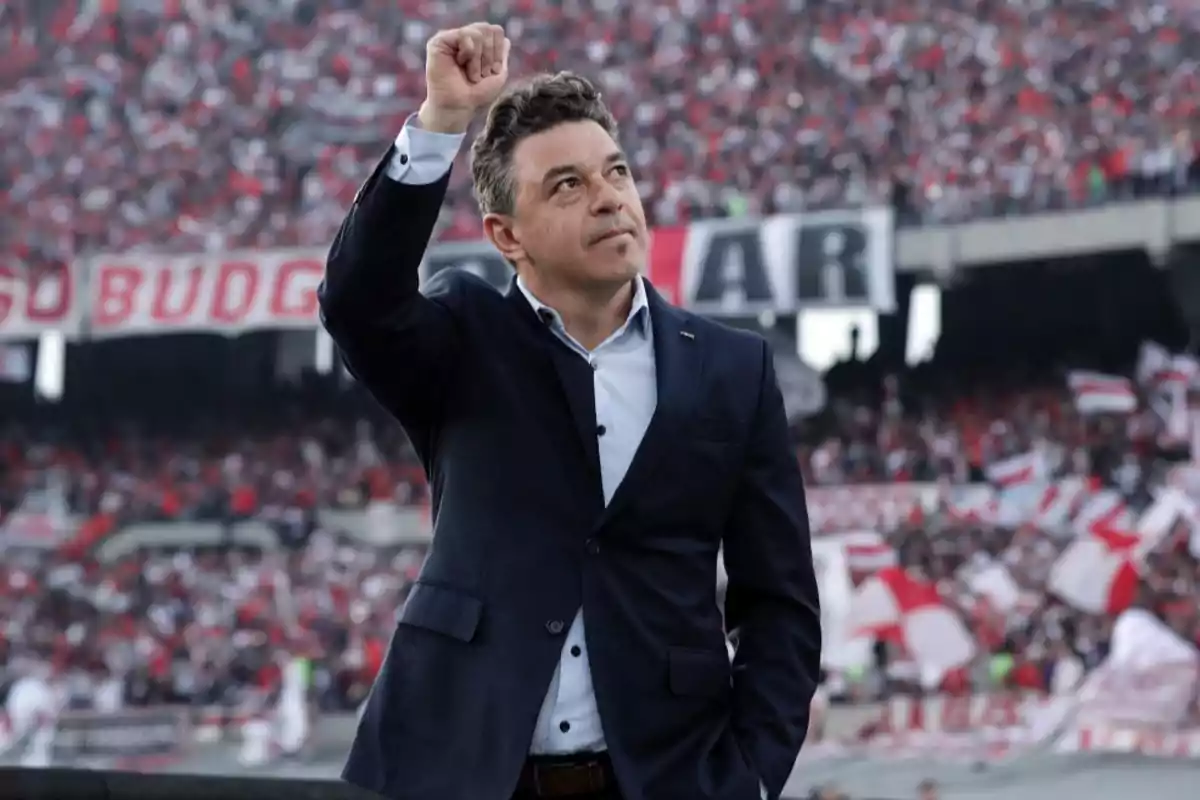 Un hombre con traje levanta el puño en un estadio lleno de aficionados con banderas rojas y blancas.