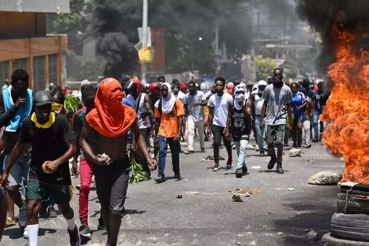 Un grupo de personas camina por una calle mientras al fondo se observa humo negro y a la derecha una barricada de neumáticos en llamas Un grupo de personas camina por una calle mientras al fondo se observa humo negro y a la derecha una barricada de neumáticos en llamas