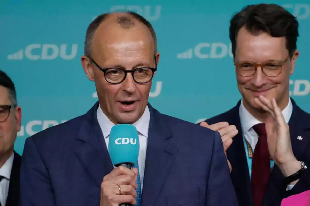 Un hombre con gafas y traje habla por un micrófono azul con el logo de CDU mientras otra persona aplaude en el fondo.