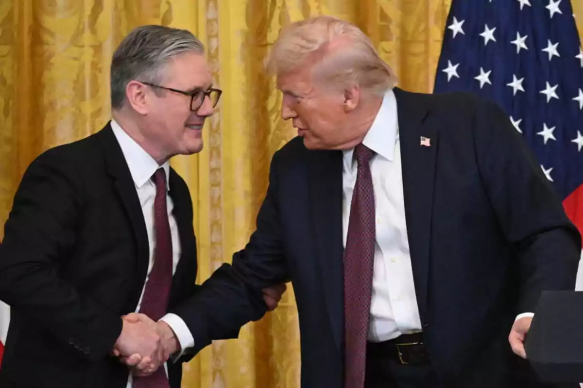 Two men in suits shake hands at a formal event with a flag in the background.