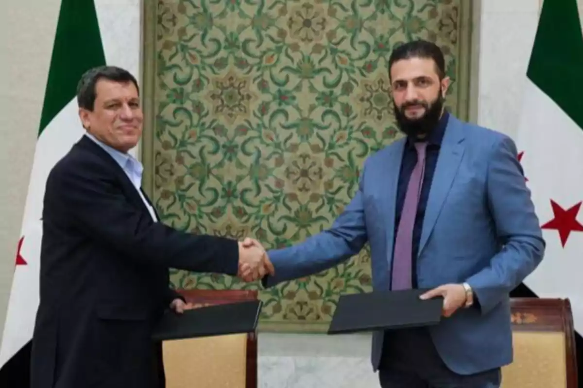 Two men standing shaking hands while holding folders, with flags and a decorative background behind them.