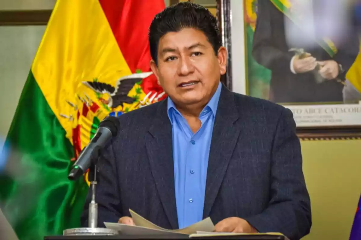 Un hombre hablando frente a un micrófono con la bandera de Bolivia detrás.