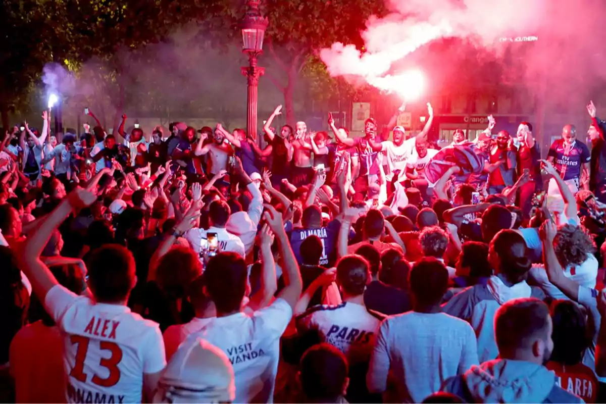 Aficionados celebrando en la calle durante la noche con bengalas y camisetas de fútbol rodeados de humo rojo
