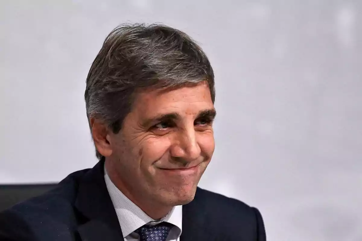 Un hombre de cabello canoso y traje oscuro sonríe ligeramente frente a un fondo gris.