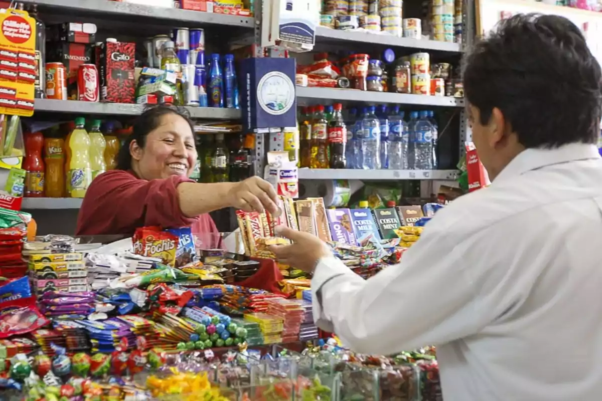 Una mujer sonriente atiende a un cliente en una tienda de abarrotes llena de productos y golosinas.