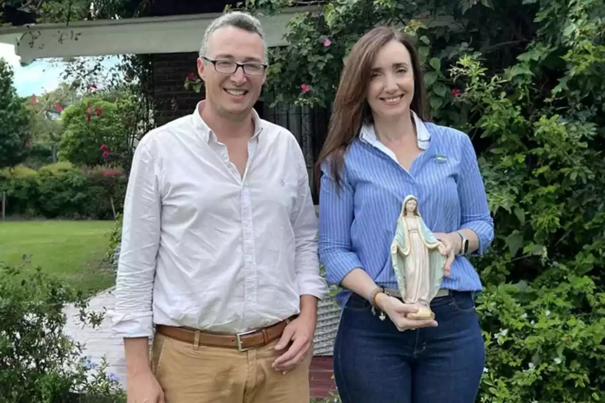 Dos personas posan en un jardín, una de ellas sostiene una estatua de la Virgen María.