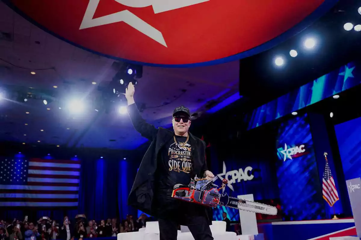 Hombre con gafas oscuras y gorra sostiene una motosierra en un escenario de conferencia con banderas de Estados Unidos y el logo de CPAC al fondo Hombre con gafas oscuras y gorra sostiene una motosierra en un escenario de conferencia con banderas de Estados Unidos y el logo de CPAC al fondo