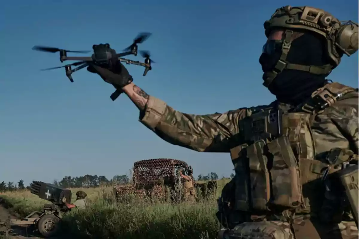 Un soldado en uniforme militar sostiene un dron en un campo abierto con vehículos camuflados al fondo.