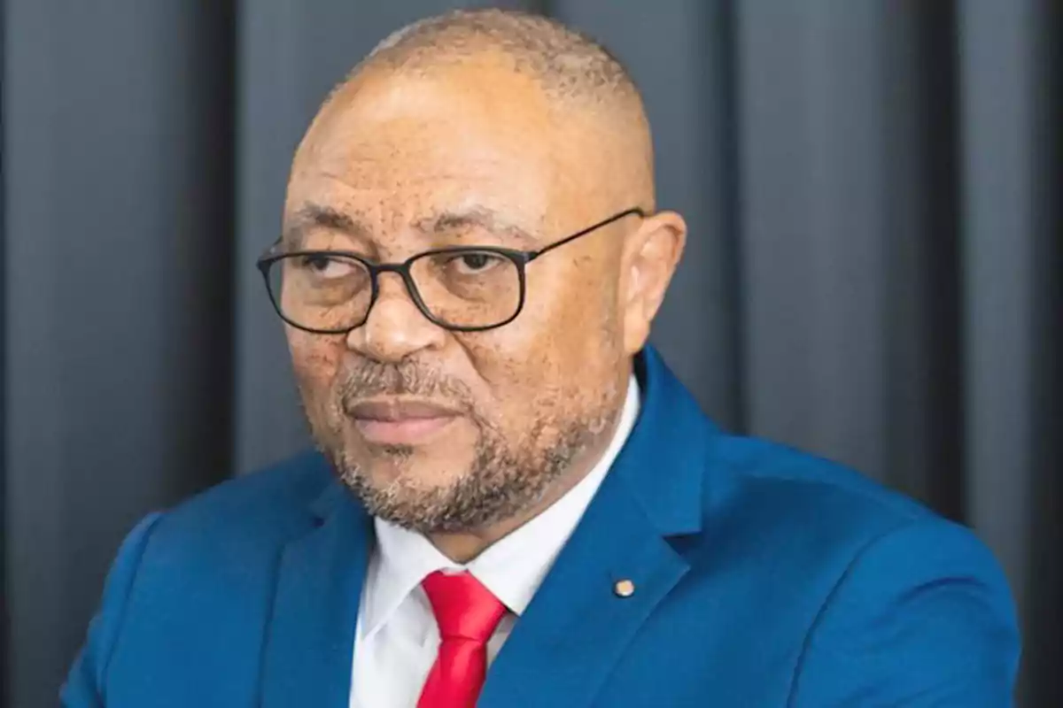 Man with glasses and a short beard wearing a blue suit and red tie in front of a dark background Man with glasses and a short beard wearing a blue suit and red tie in front of a dark background