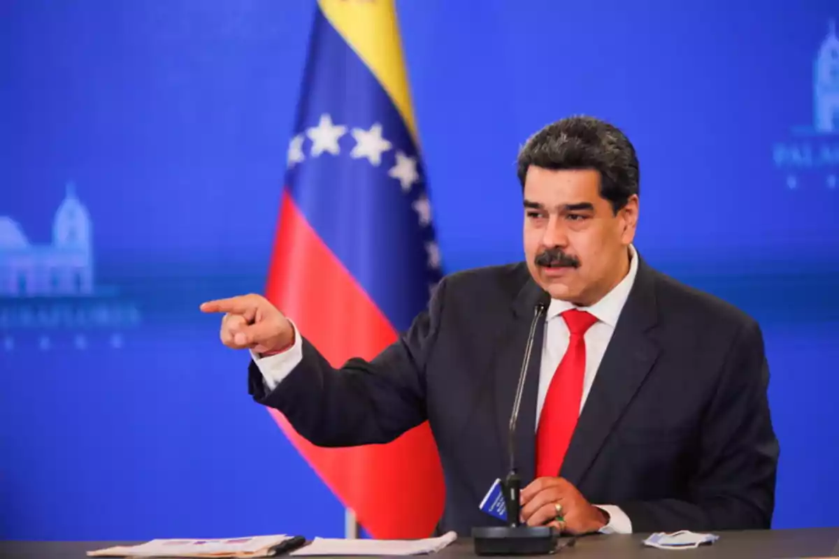 Un hombre en traje oscuro y corbata roja habla en un podio con un micrófono, señalando con el dedo, frente a una bandera de Venezuela.