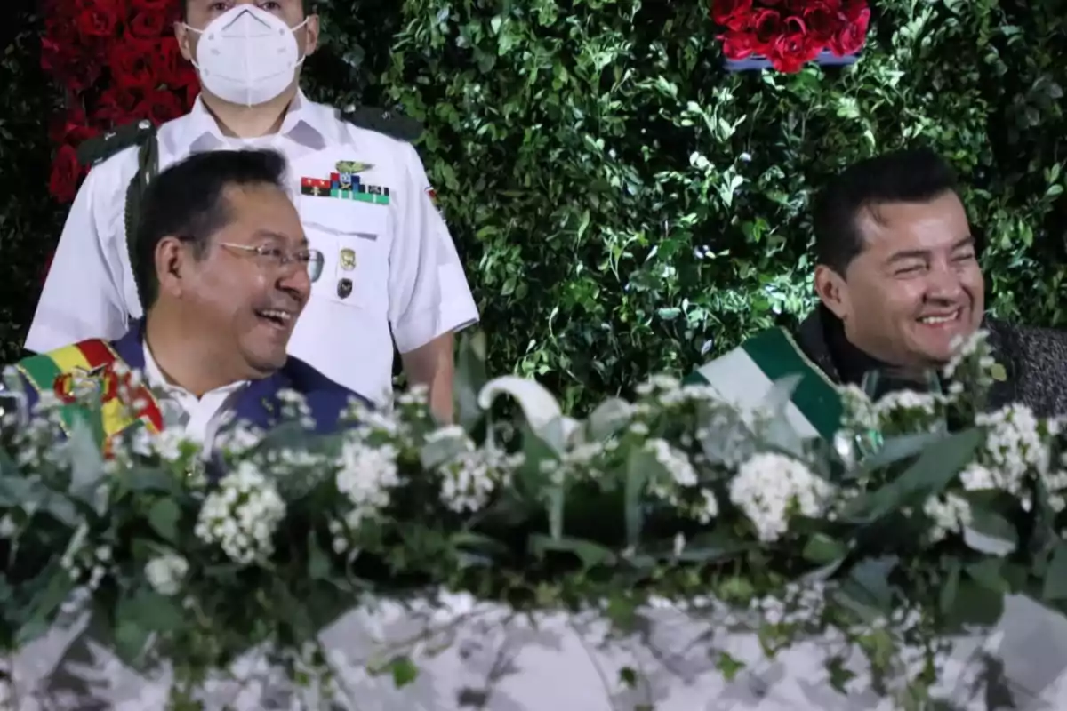 Two men smile while sitting behind a table decorated with white and green flowers, as a man in a white uniform and mask stands behind them, with a background of foliage and red roses. Two men smile while sitting behind a table decorated with white and green flowers, as a man in a white uniform and mask stands behind them, with a background of foliage and red roses.
