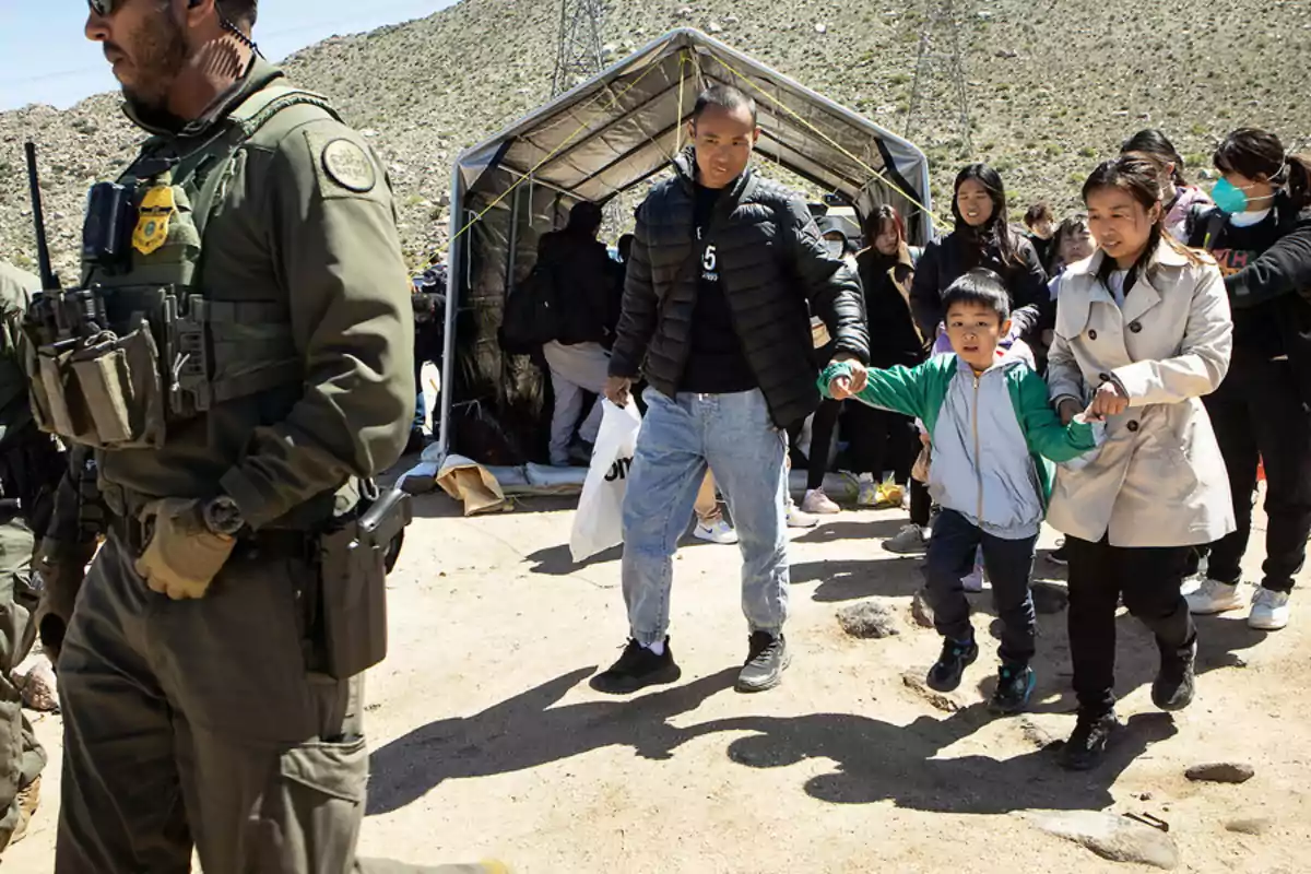 Un grupo de personas, incluyendo un niño, camina de la mano junto a un oficial de seguridad en un entorno al aire libre con una tienda de campaña en el fondo.