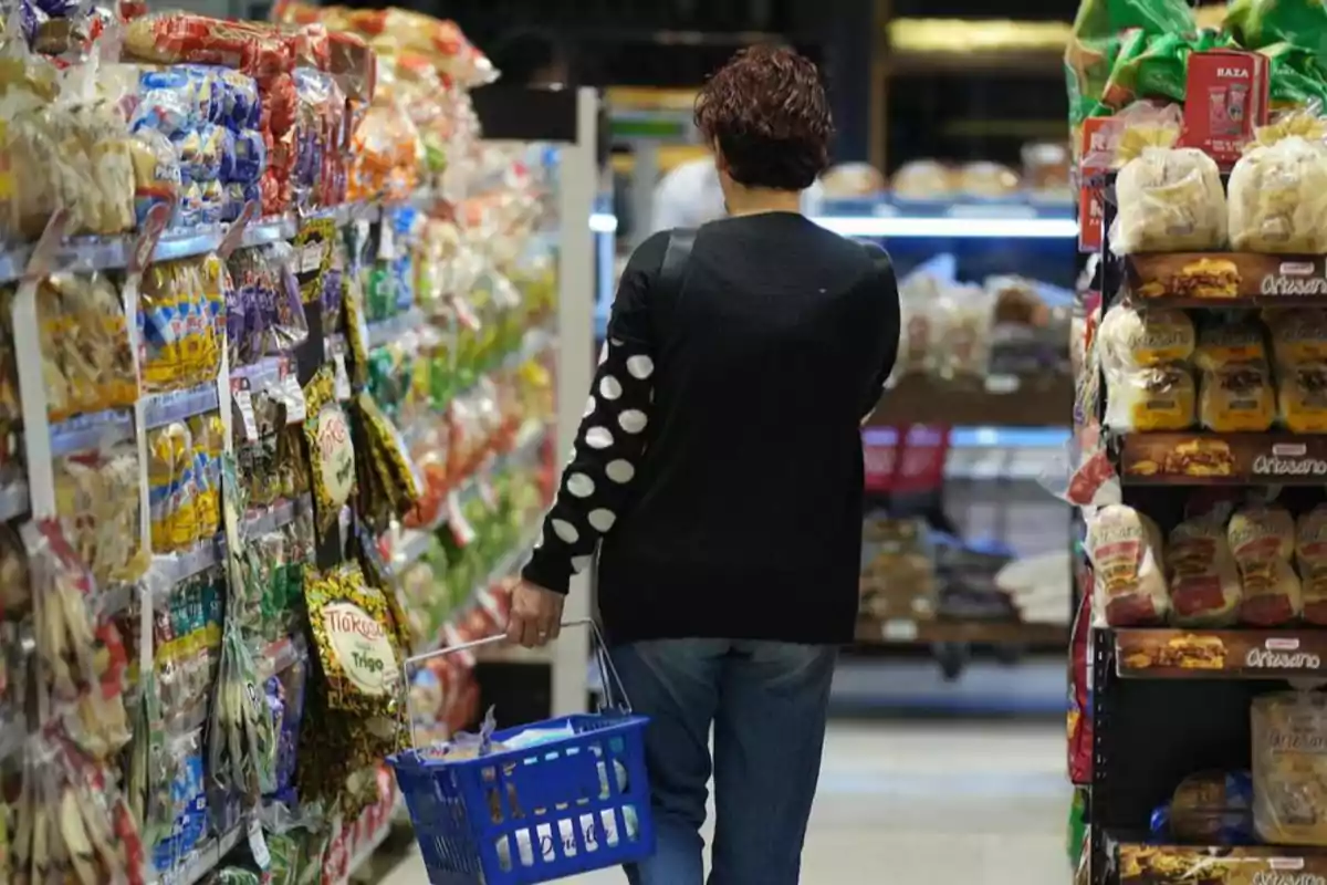 Persona de espaldas con suéter negro y puntos blancos en la manga caminando por un pasillo de supermercado con una canasta azul en la mano rodeada de estantes llenos de productos empaquetados Persona de espaldas con suéter negro y puntos blancos en la manga caminando por un pasillo de supermercado con una canasta azul en la mano rodeada de estantes llenos de productos empaquetados