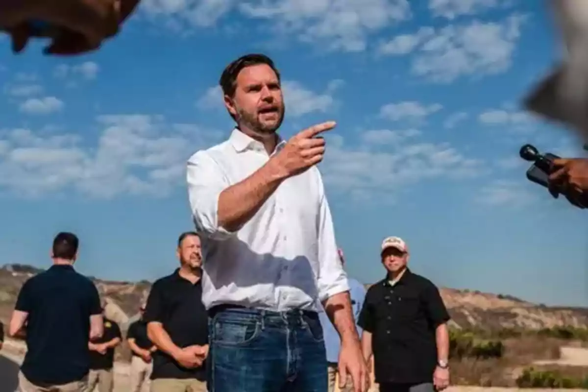 Hombre de pie al aire libre con camisa blanca y jeans rodeado de personas y micrófonos bajo un cielo azul con nubes.