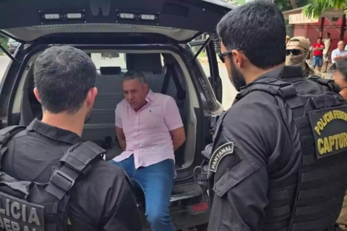 Un hombre con camisa rosa está sentado en la parte trasera de un vehículo con las manos atadas mientras es custodiado por agentes de policía vestidos de negro.