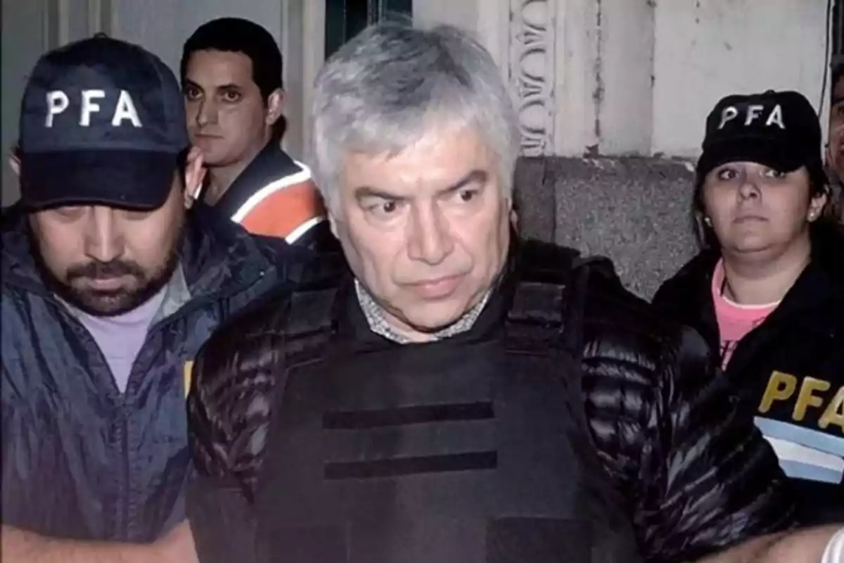 Un hombre de cabello canoso es escoltado por agentes de la policía federal argentina que llevan gorras con las siglas PFA