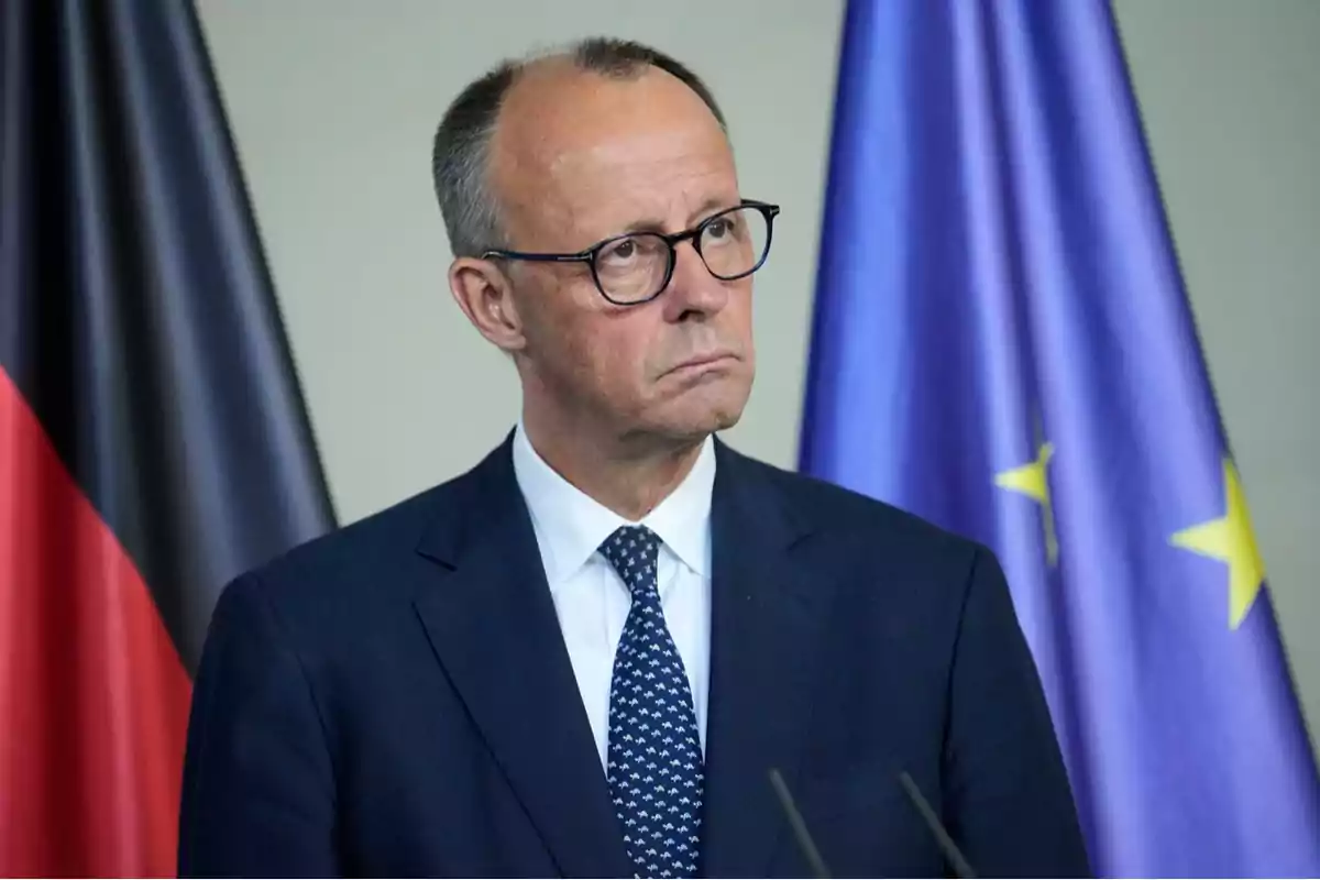 A man with glasses and a dark suit is standing in front of two flags, one of Germany and the other of the European Union. A man with glasses and a dark suit is standing in front of two flags, one of Germany and the other of the European Union.