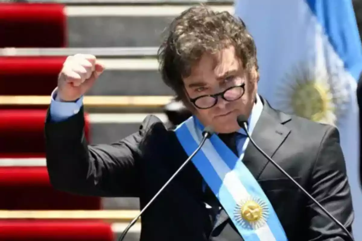 Hombre con gafas y traje oscuro, usando una banda presidencial azul y blanca, levanta el puño mientras habla en un podio con micrófonos, con una bandera de fondo.