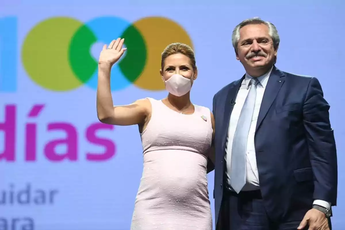 Una mujer embarazada con mascarilla y un hombre de traje sonríen y posan frente a un fondo con texto colorido.