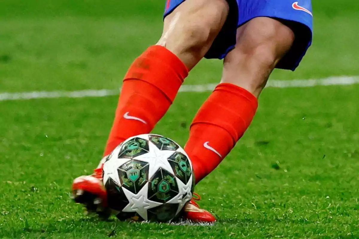 Piernas de un futbolista con medias rojas y shorts azules pateando un balón de fútbol sobre césped Piernas de un futbolista con medias rojas y shorts azules pateando un balón de fútbol sobre césped