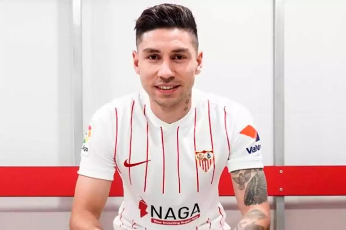 Un jugador de fútbol con la camiseta del Sevilla FC sonríe mientras está sentado en un vestuario.