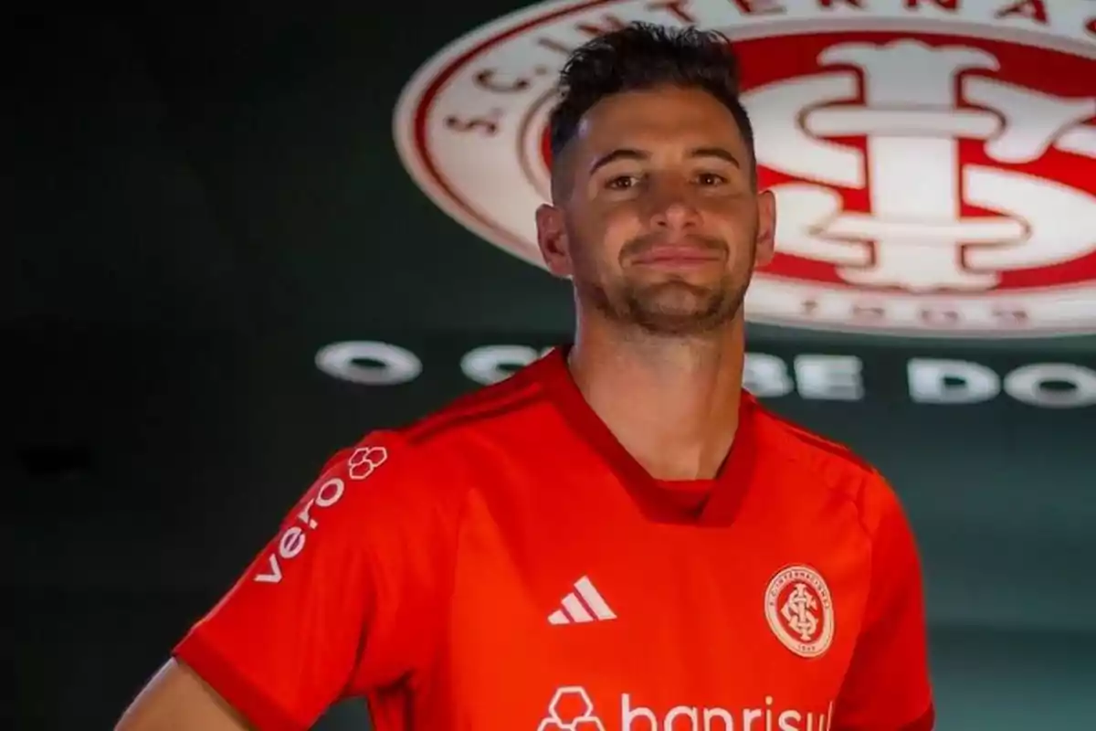 Un jugador de fútbol con camiseta roja del S.C. Internacional posa frente al escudo del club.