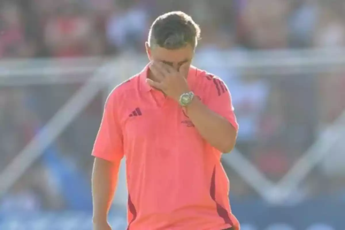 Un hombre con una camiseta rosa se cubre el rostro con la mano en un campo deportivo.