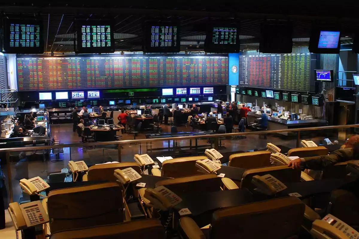 Sala de operaciones de una bolsa de valores con múltiples pantallas mostrando datos financieros y personas trabajando en escritorios.