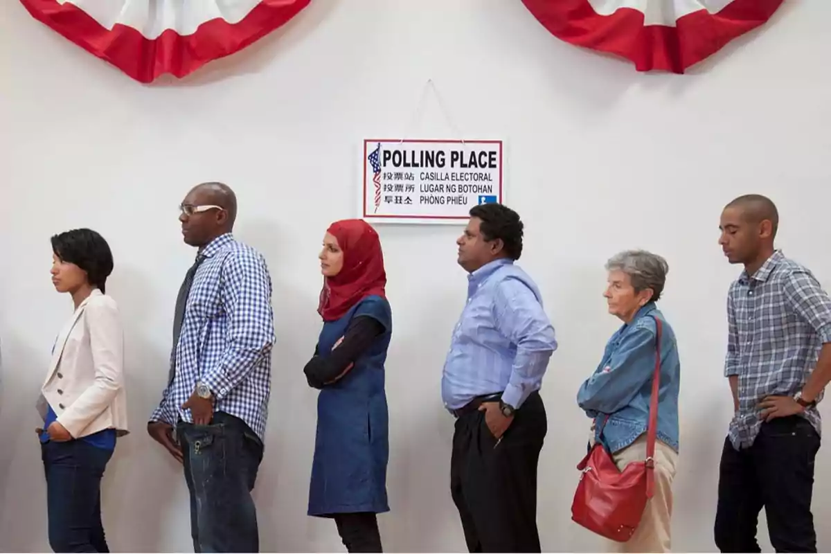 Personas de pie en fila en un lugar de votación con un cartel en la pared y una decoración de tela roja y blanca arriba.