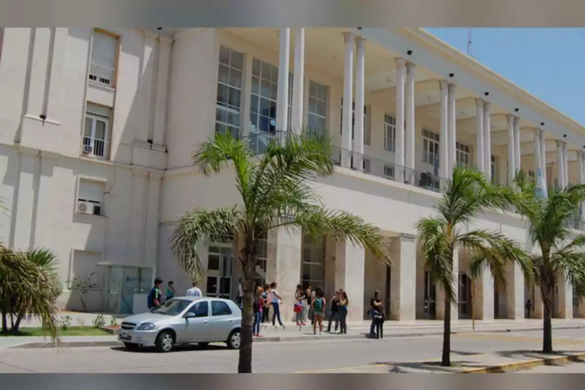 Fachada de un edificio grande con columnas blancas, palmeras al frente, un auto plateado estacionado y varias personas reunidas en la entrada