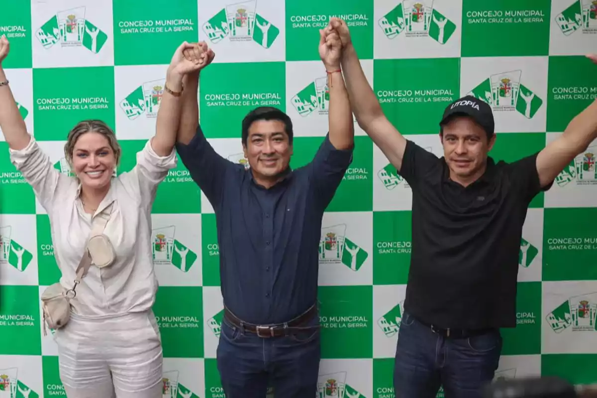 Three smiling people raise their joined hands in front of a background with the logo of the Santa Cruz de la Sierra City Council. Three smiling people raise their joined hands in front of a background with the logo of the Santa Cruz de la Sierra City Council.
