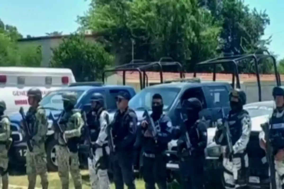 Un grupo de personas uniformadas y armadas está de pie frente a varios vehículos, incluyendo una ambulancia, en un entorno al aire libre con árboles al fondo.